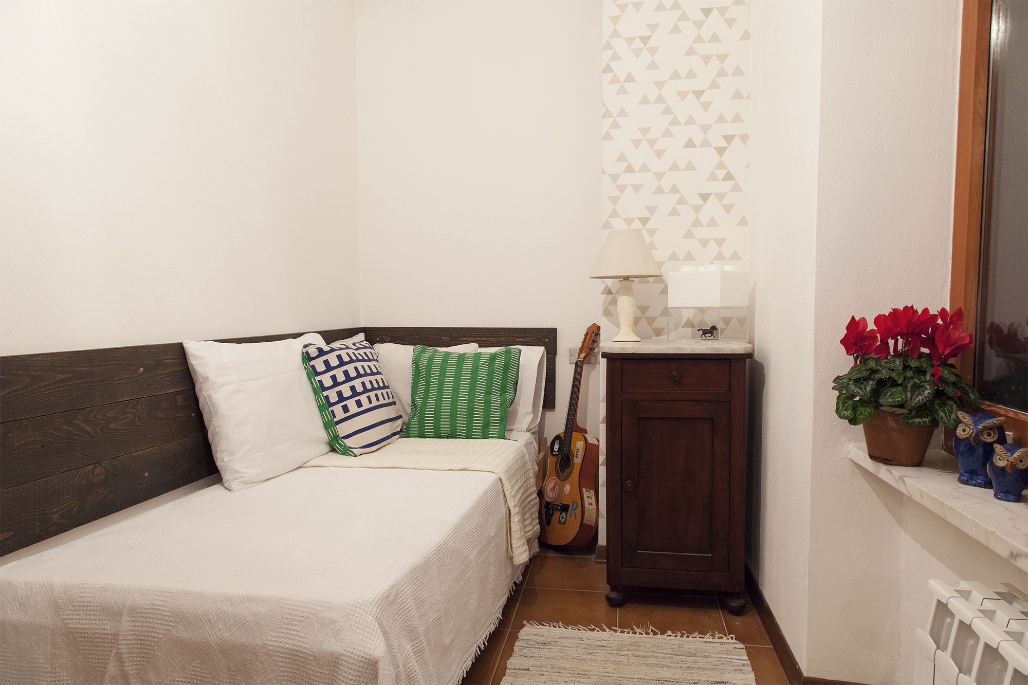 A small, cozy bedroom with a bed, a guitar, a lamp, and a window with a view of the outside.