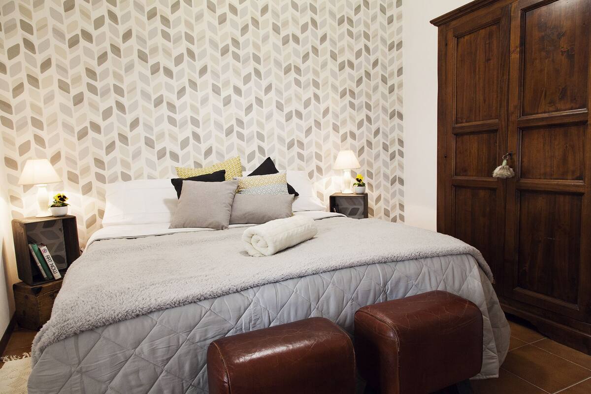 A neatly arranged bedroom with a bed, two nightstands, a wardrobe, and a pair of leather ottomans.