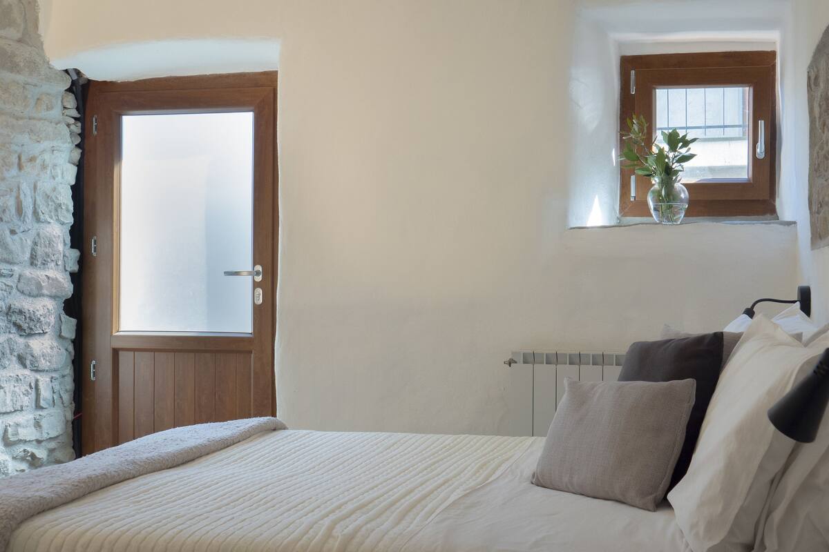 A bedroom with a stone wall, a wooden door, a window, a bedside table with a vase, and a bed with pillows and a blanket.