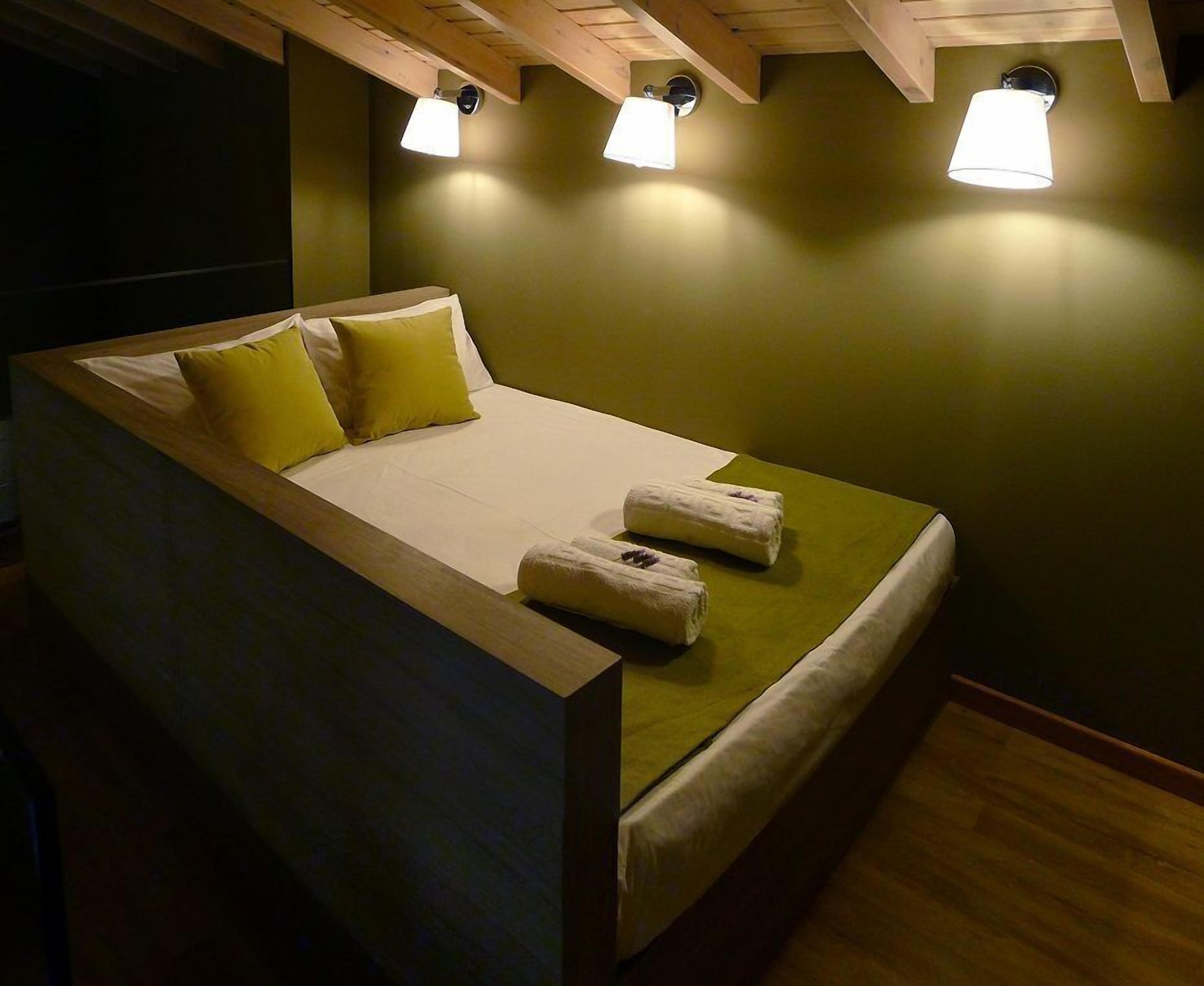 A single bed with a wooden headboard, two pillows, and two towels on a green mattress in a room with wooden beams and three wall-mounted lights.
