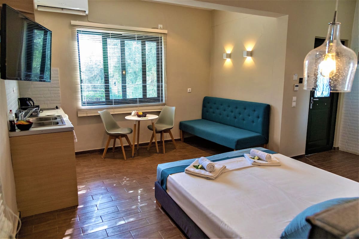 Habitación de hotel con cama, sofá, pequeña mesa con sillas, zona de cocina y ventana con persianas.