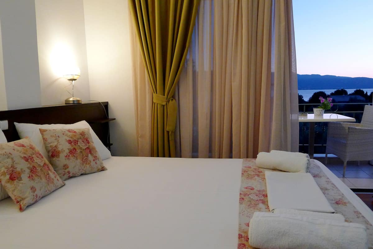 Habitación de hotel con cama, almohadillas florales, lámpara de noche, vista al mar y balcón con silla y mesa.