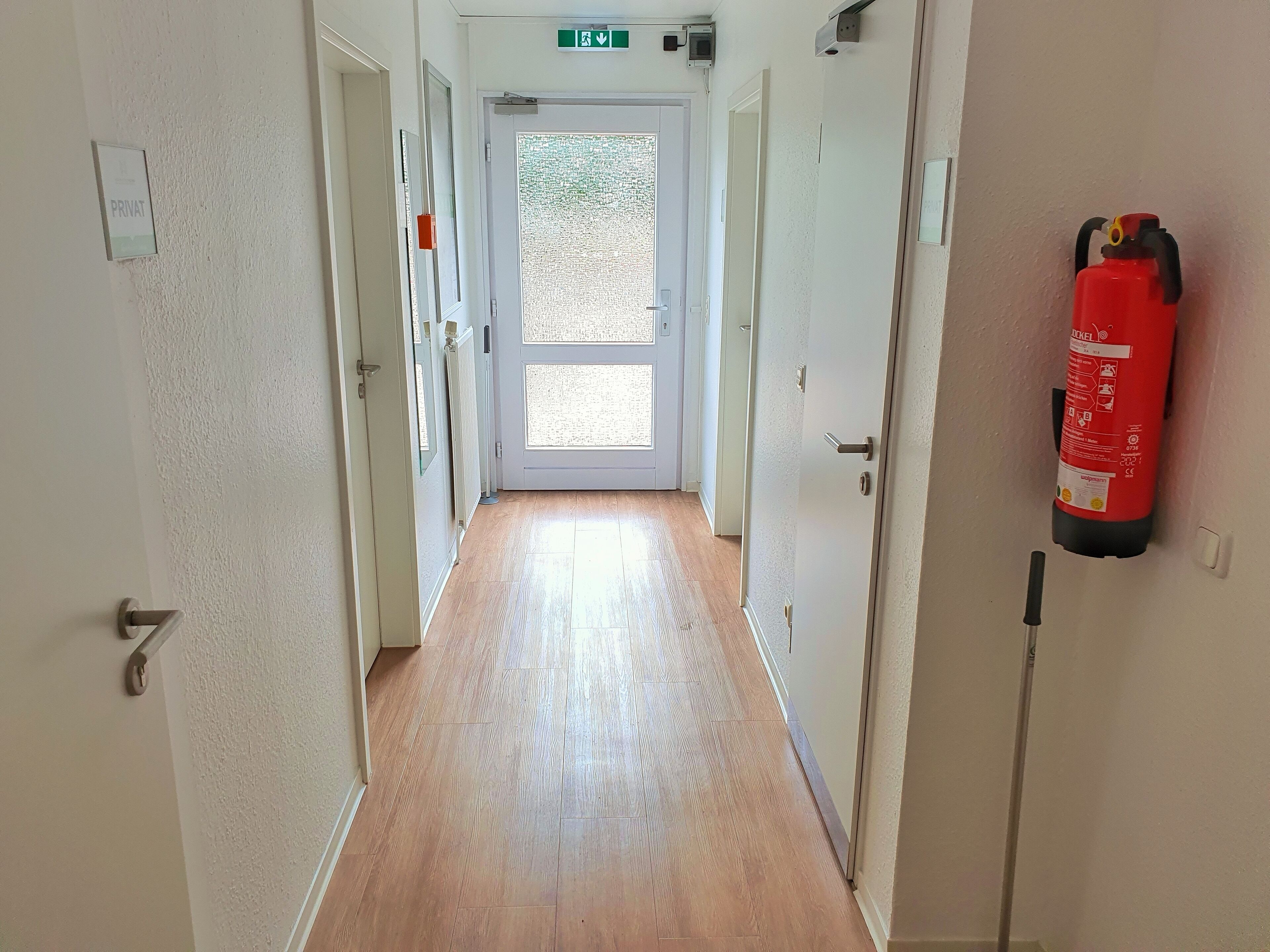 Un couloir avec un extincteur fixé au mur, une porte au fond et une pancarte au-dessus de la porte.
