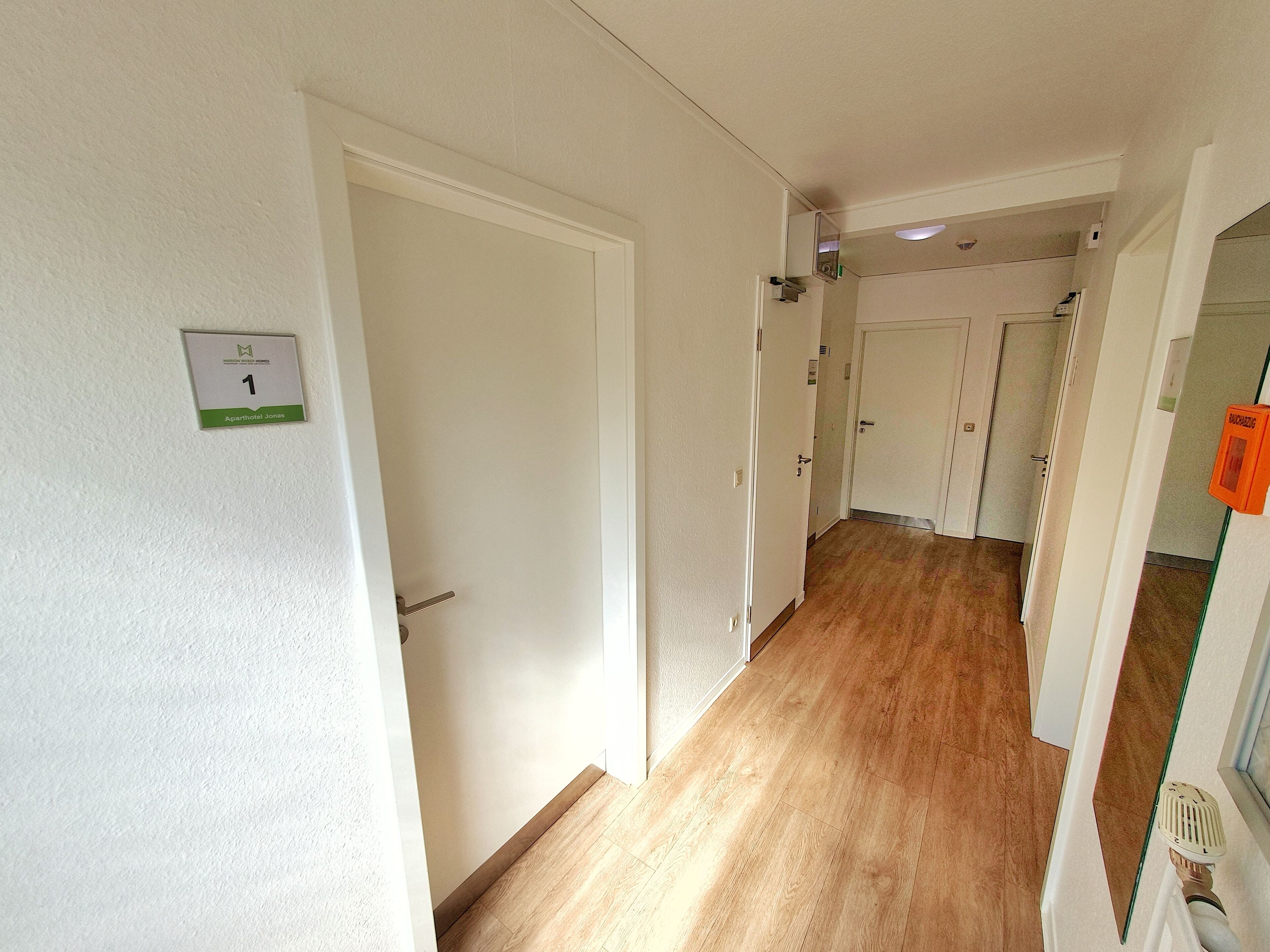 A hallway with a wooden floor, white walls, and doors leading to rooms. A sign indicates room number 1.