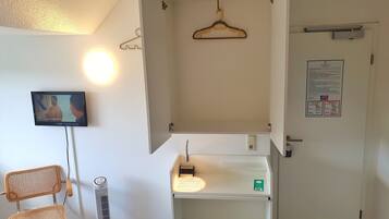 A room with a television mounted on the wall, a chair, a standing fan, a white cabinet with a mirror, and a door with a notice on it.