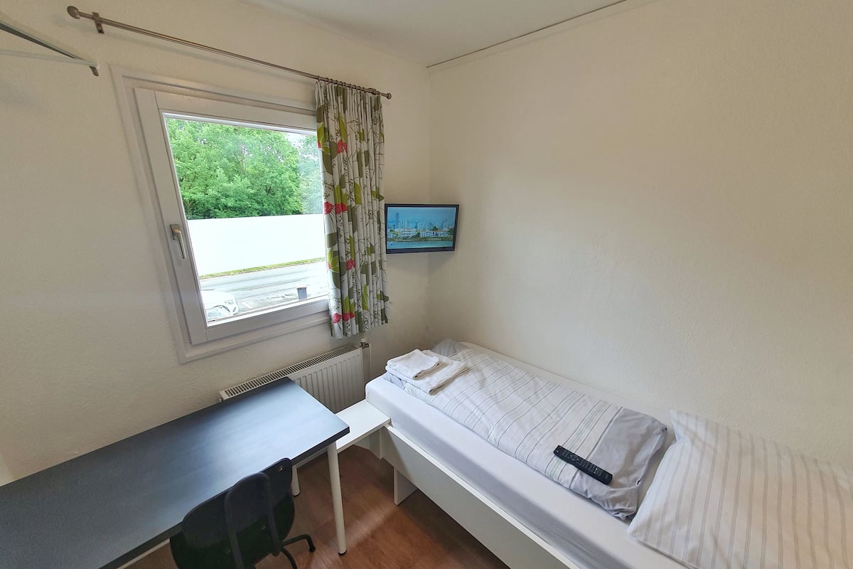 A small, tidy room with a single bed, a desk, and a chair. A window with curtains is visible, and a television is mounted on the wall.