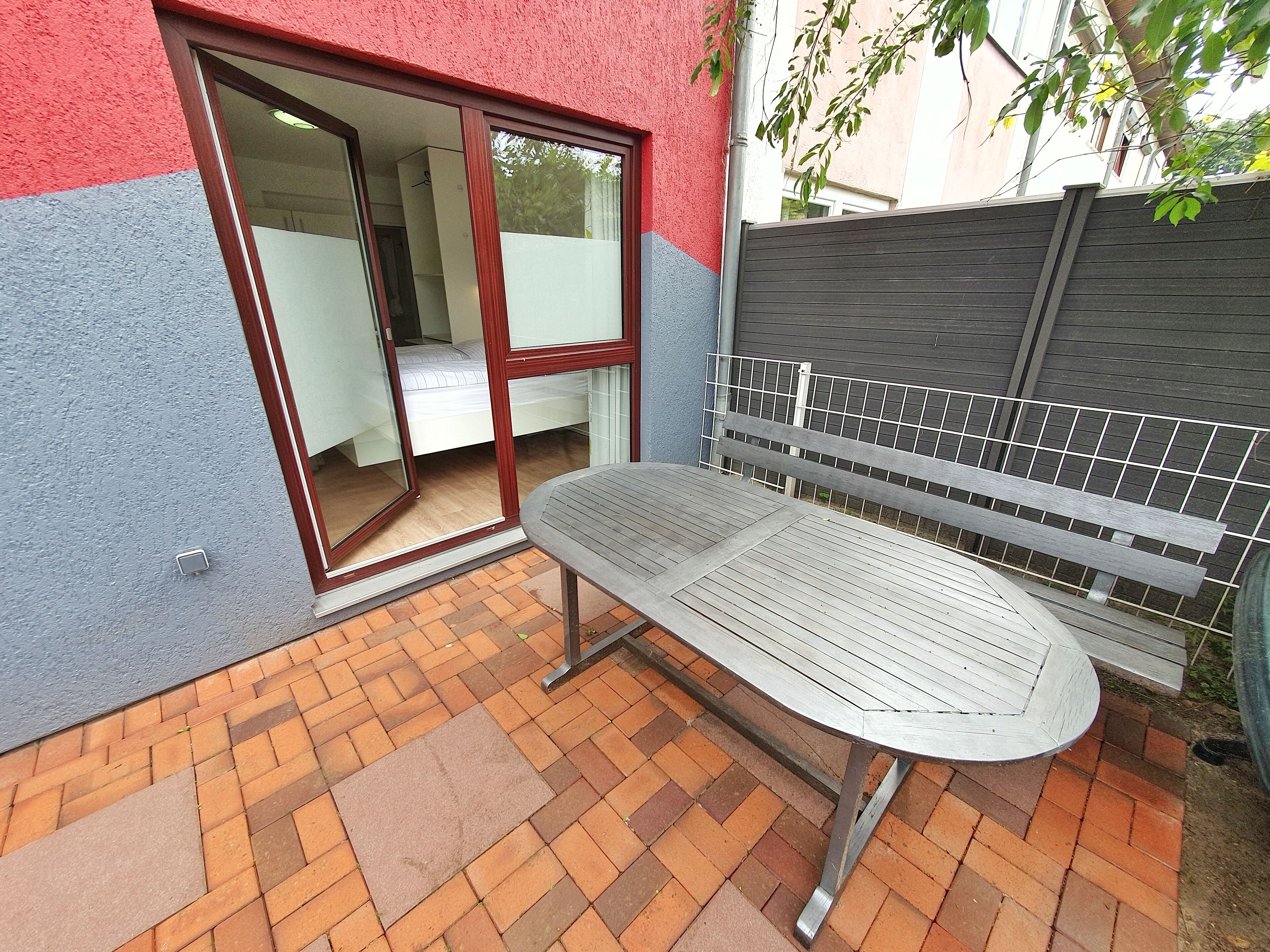 A patio with a metal table and bench, a sliding glass door leading to a bedroom, and a grey fence.