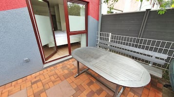 A patio with a metal table and bench, a sliding glass door leading to a bedroom, and a grey fence.