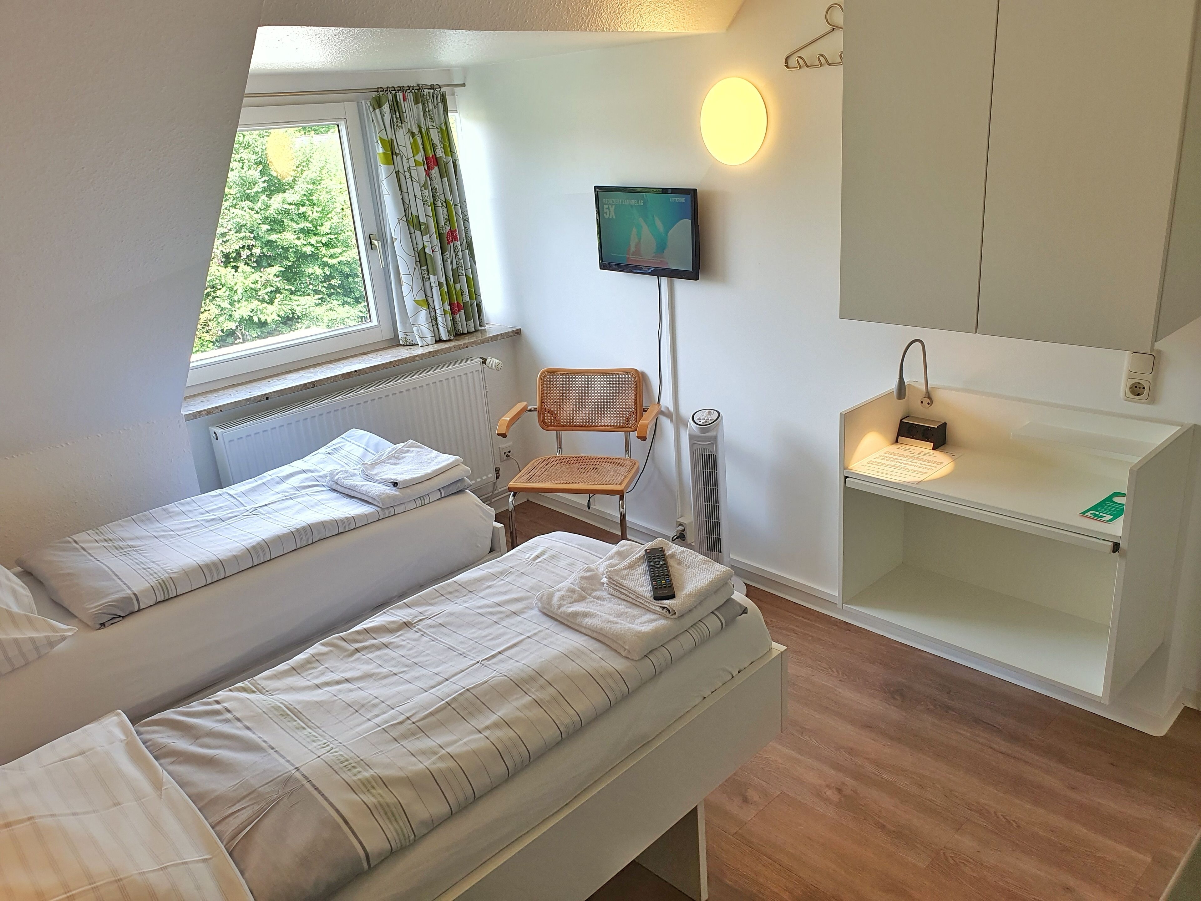 Een kleine, goed verlichte kamer met twee bedden, een stoel, een televisie en een bureau met een lamp.