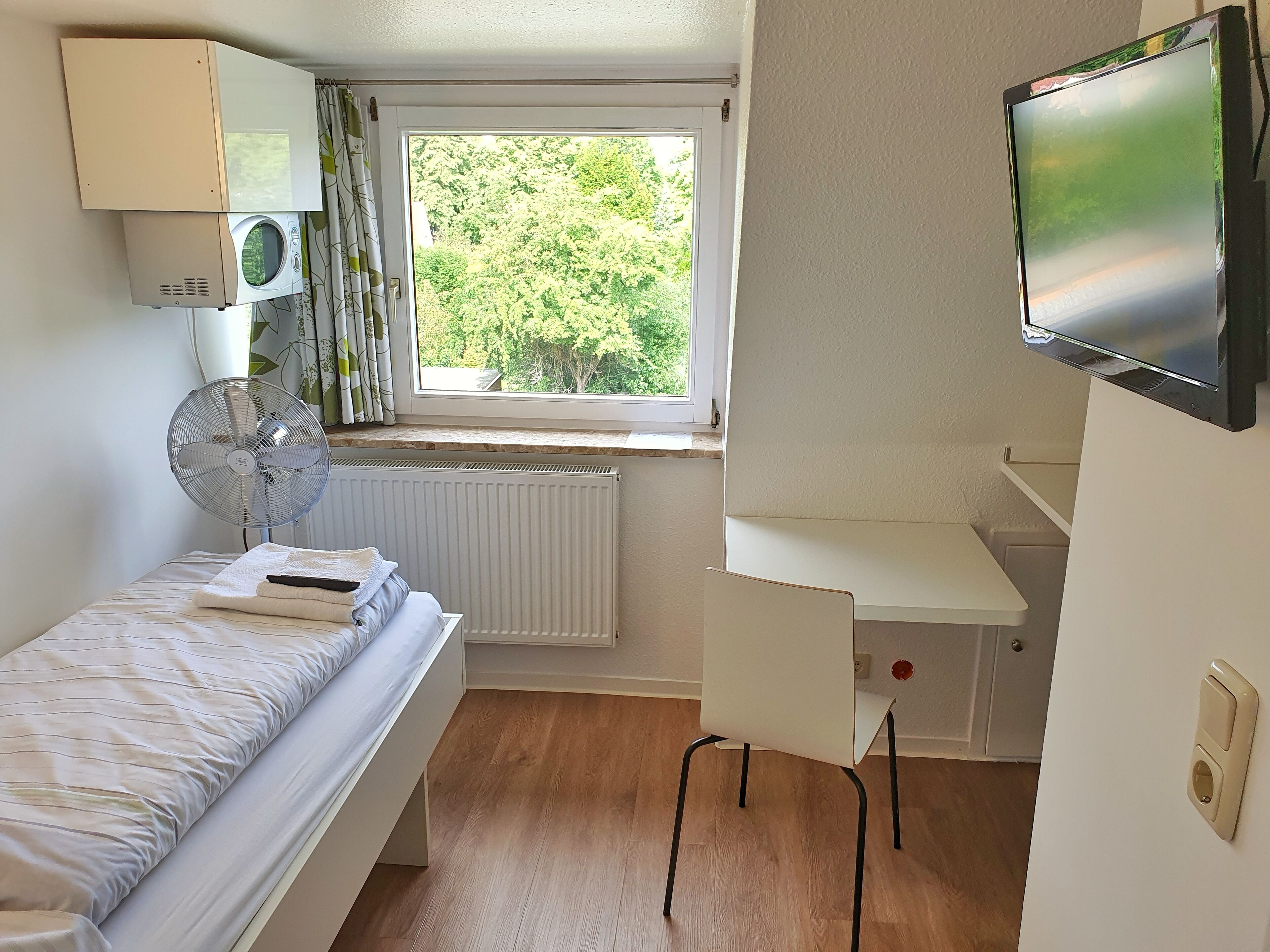 Ein Schlafzimmer mit Bett, Schreibtisch, Stuhl, Fernseher, Heizkörper und einem Fenster mit Blick auf Bäume.