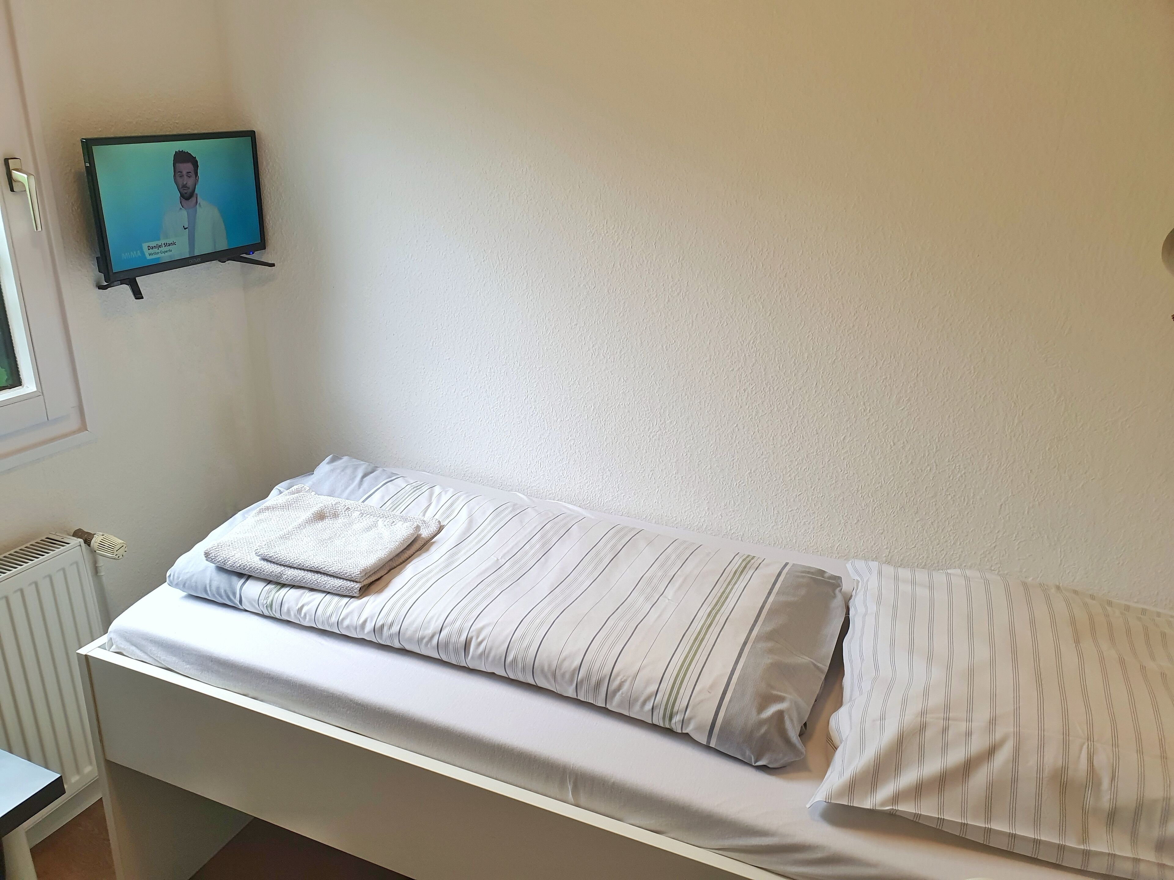 A single bed with striped bedding, a small bedside table, and a wall-mounted TV displaying a person.