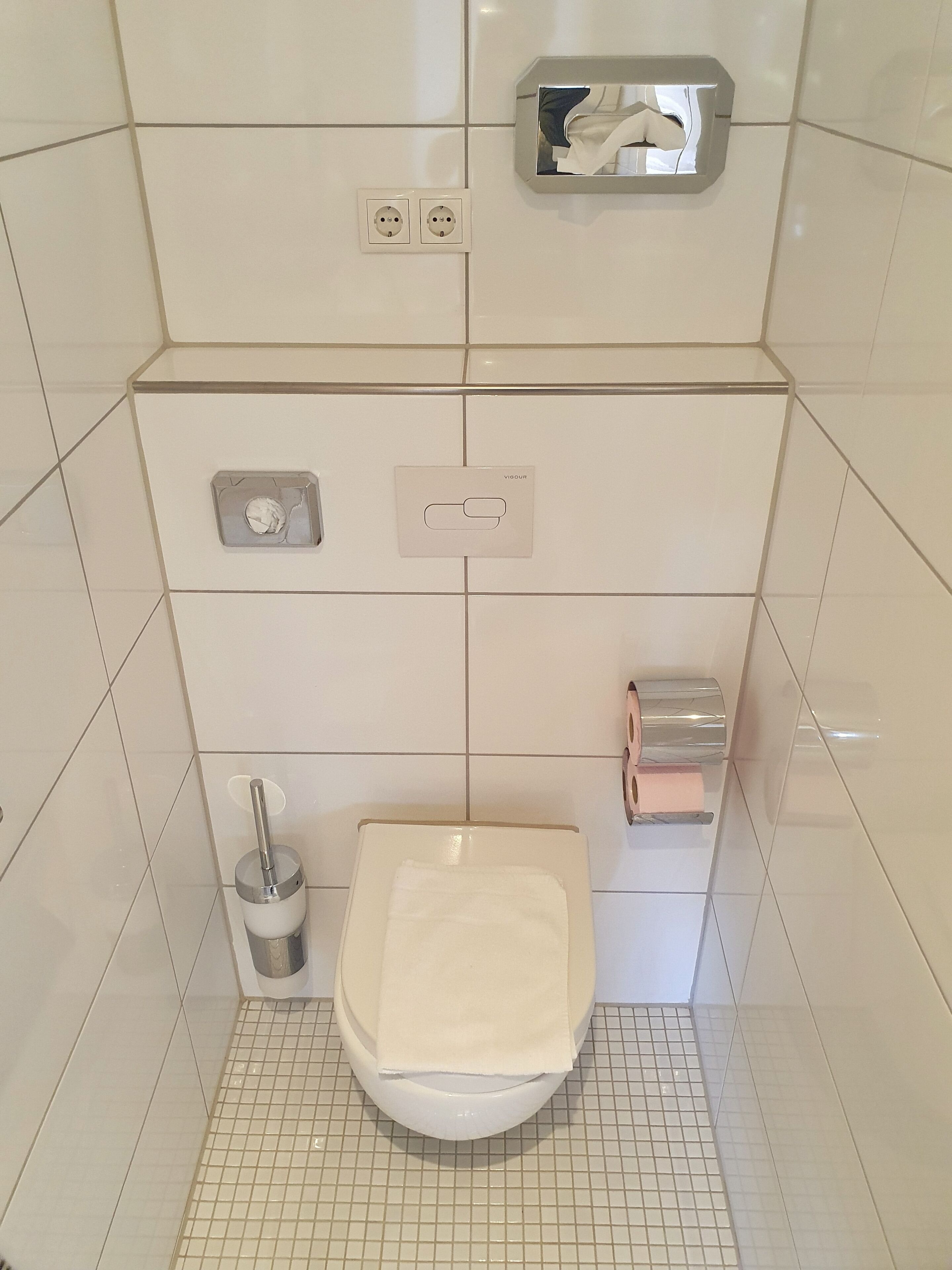 Een kleine betegelde badkamer met toilet, handdroger en papieren handdoekdispenser.