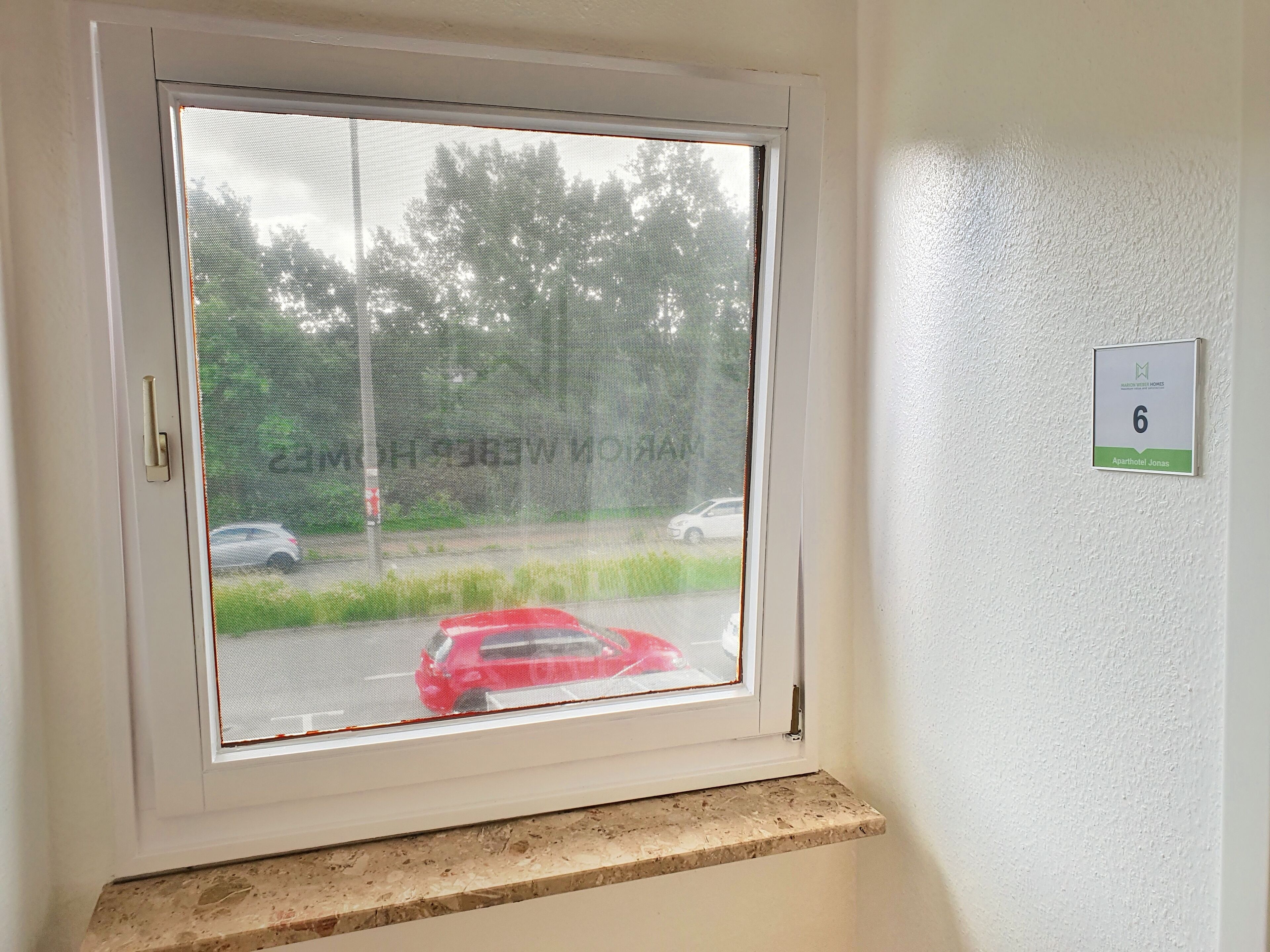 A window with a view of a road and greenery, a door handle, and a room number sign.