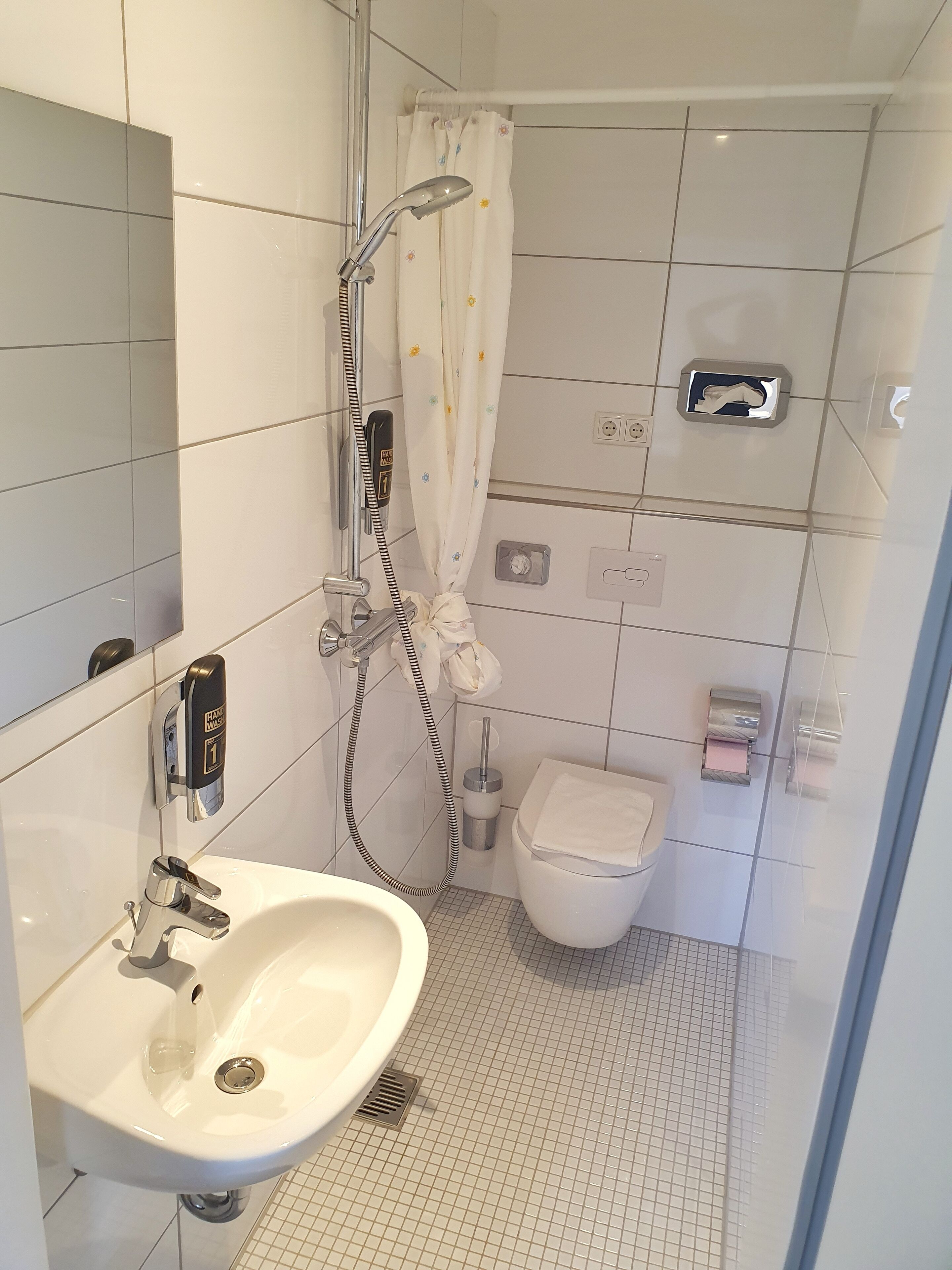 A modern bathroom with a white sink, a handheld showerhead, a toilet, and a hand dryer.