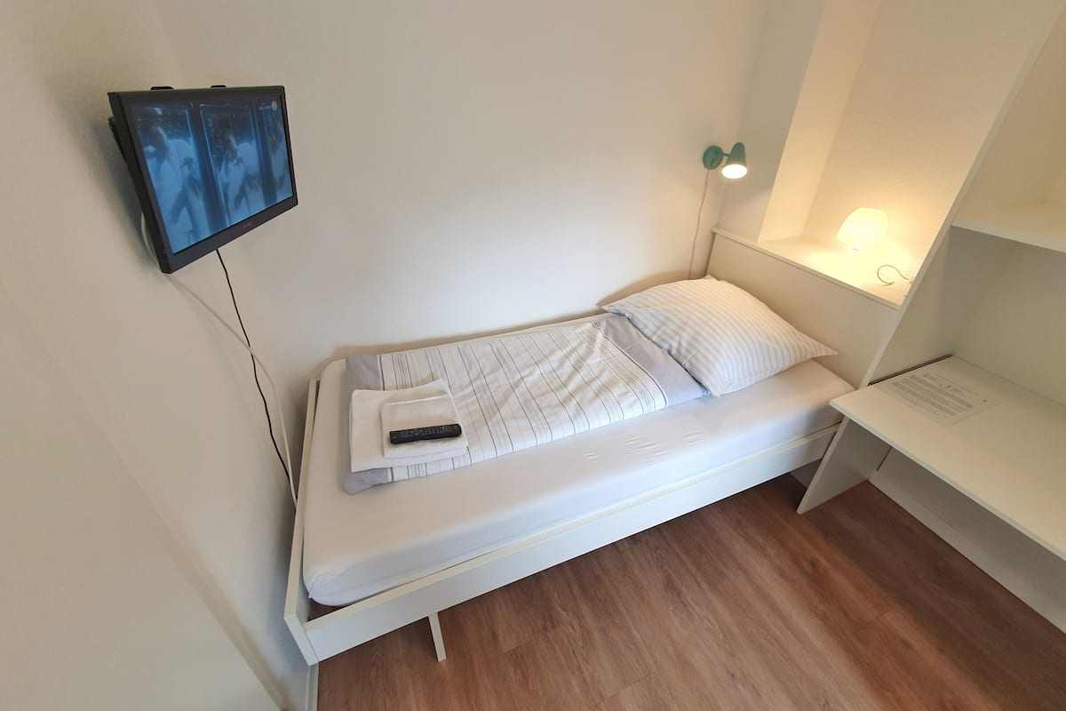A small, white, single-bed room with a television mounted on the wall, a bedside table with a lamp, and a wooden floor.