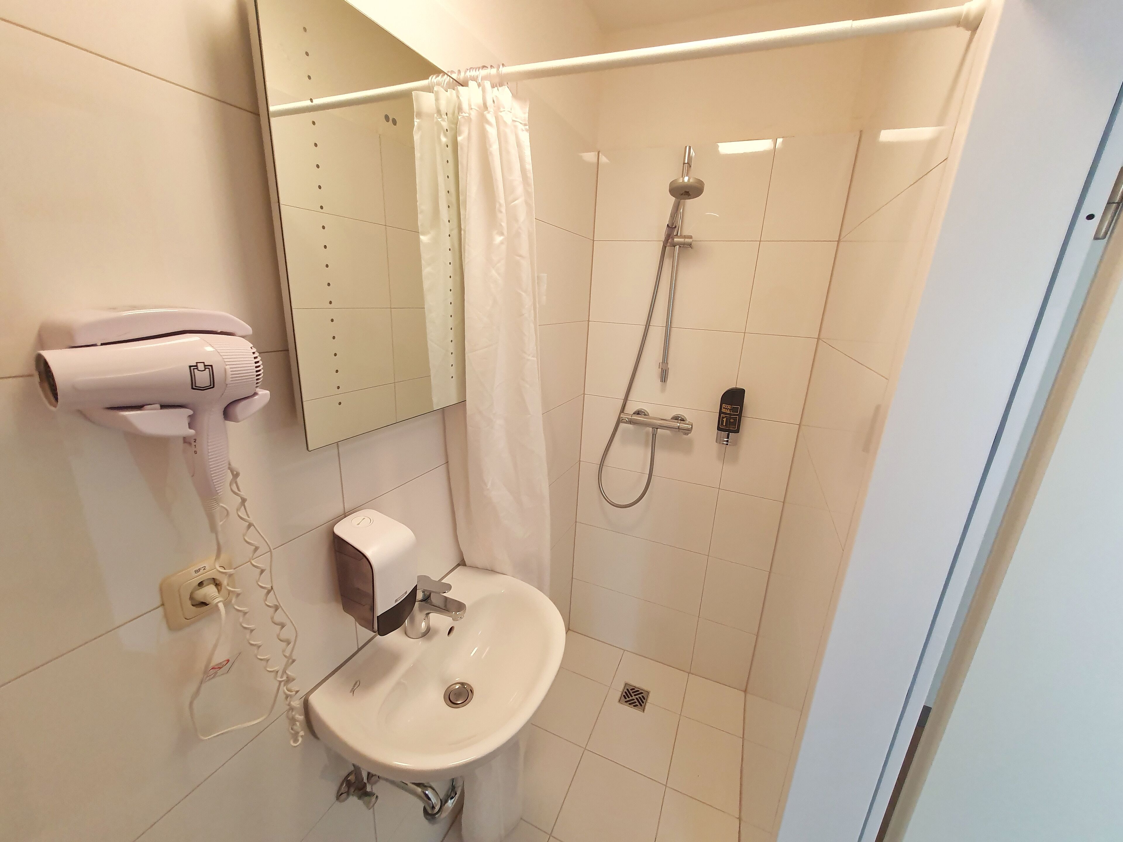 Ein Badezimmer mit einem weißen Waschbecken, einem Haartrockner, einer Dusche inklusive Duschbrause und einem Handtuchhalter.