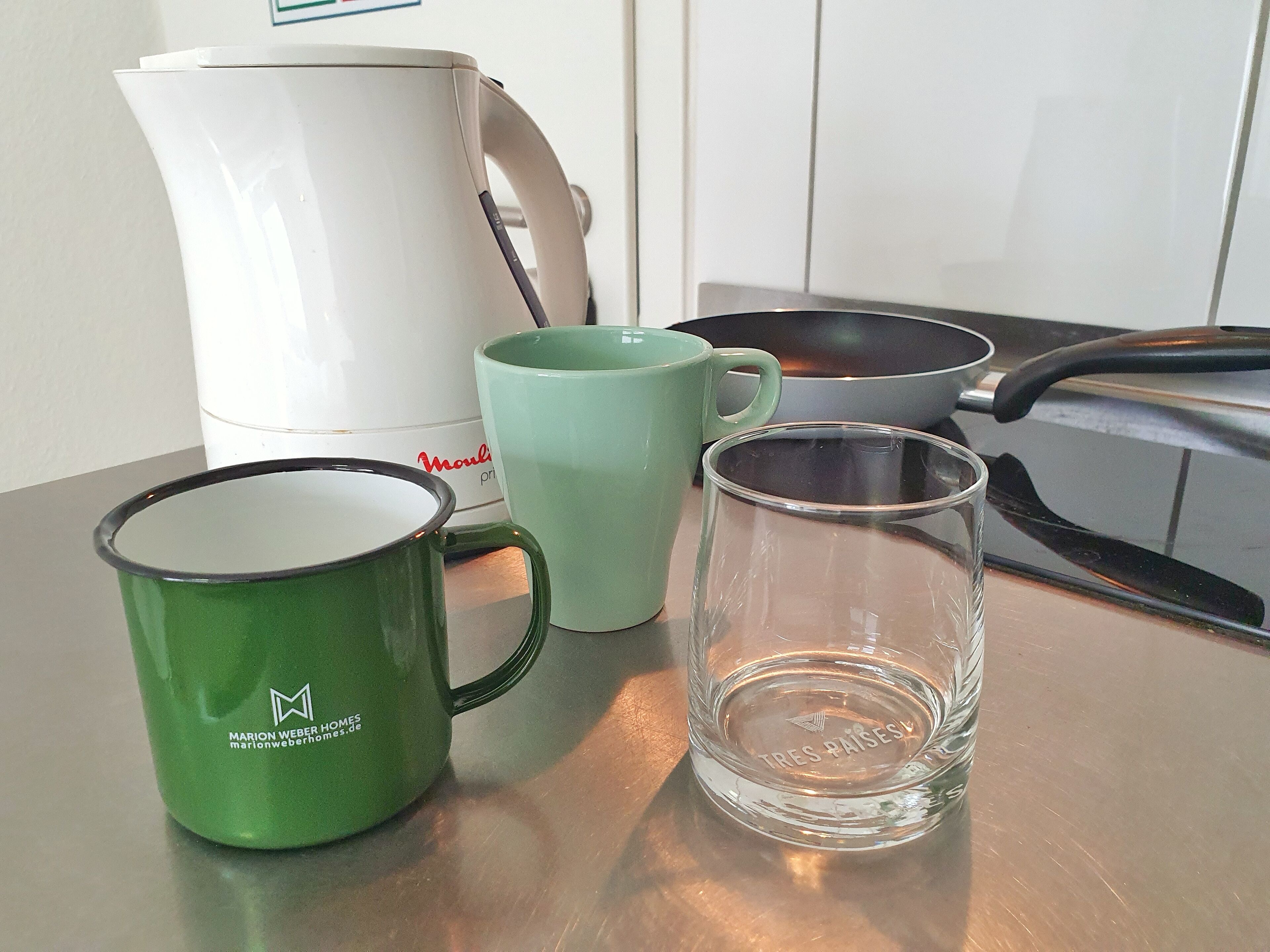 Un comptoir de cuisine avec une machine à café blanche, une tasse verte, un verre transparent et une poêle noire.