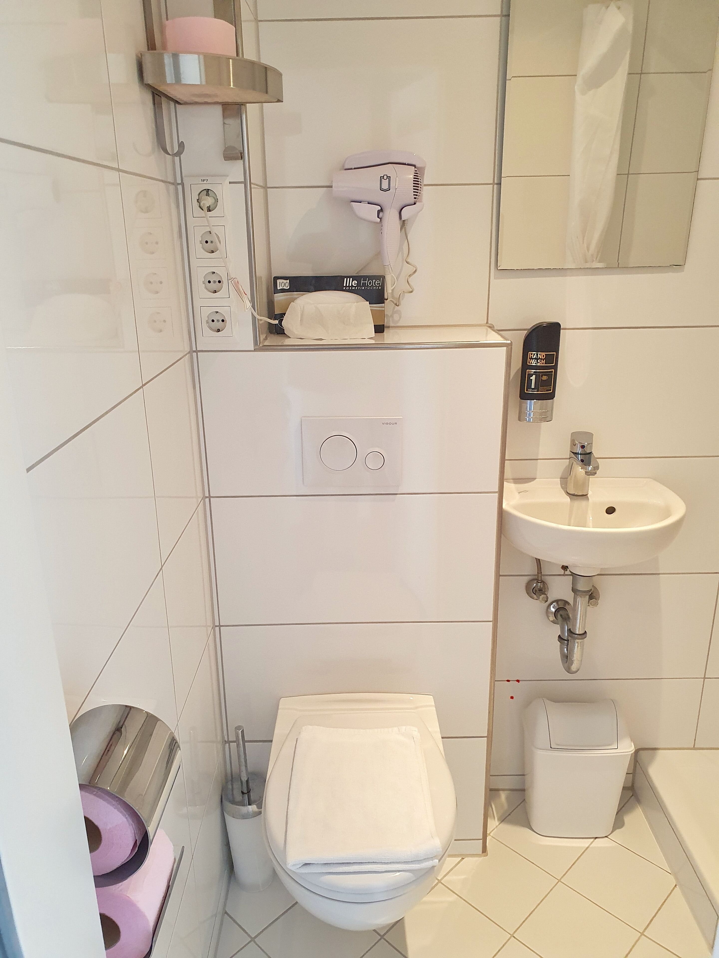 Une salle de bain équipée de toilettes, d’un lavabo et d’une douche. Un sèche-cheveux est fixé au mur, et un distributeur de gel hydroalcoolique est également présent.