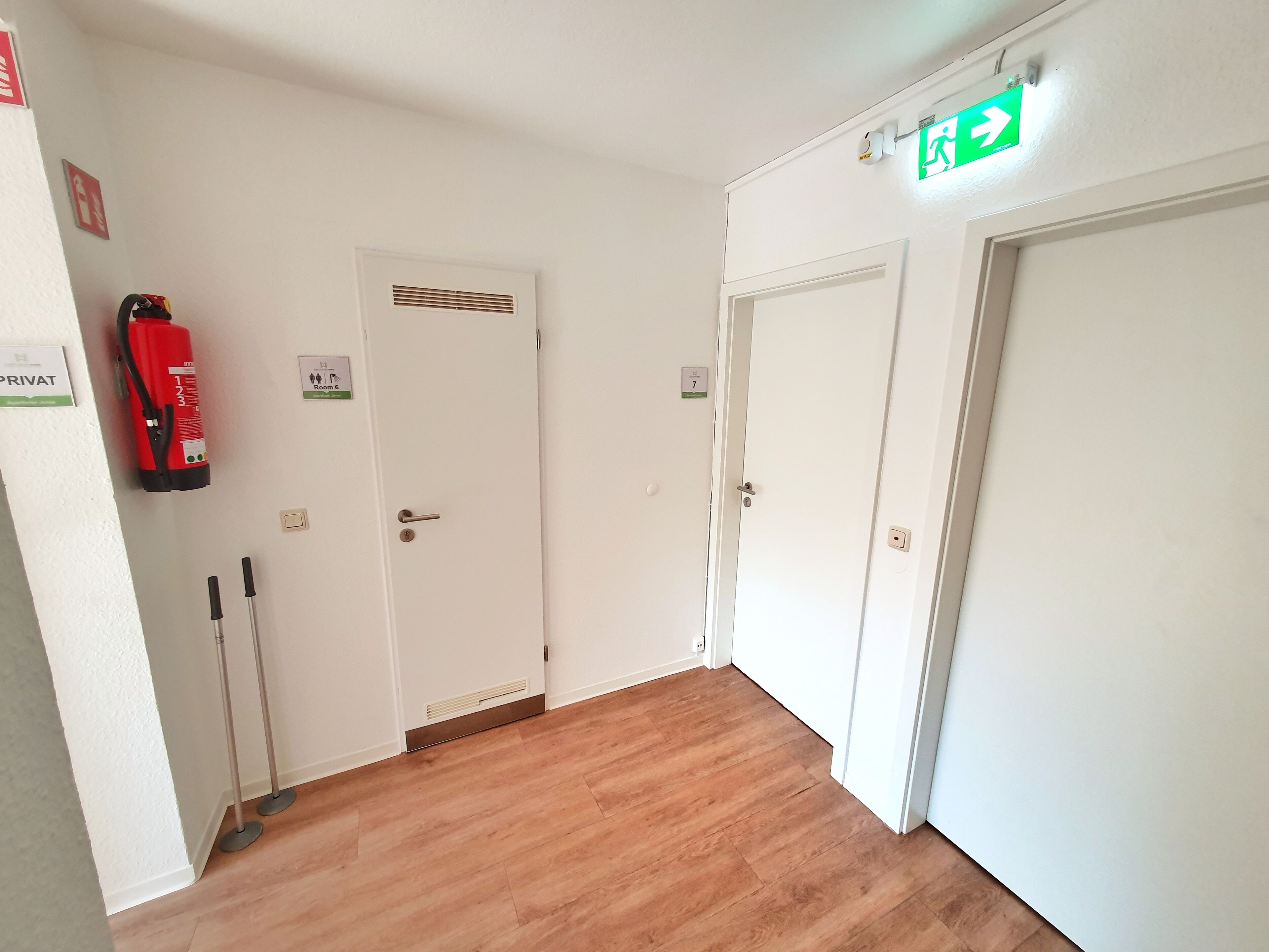 En hall med två vita dörrar, en brandsläckare och en skylt som visar var nödutgången finns.