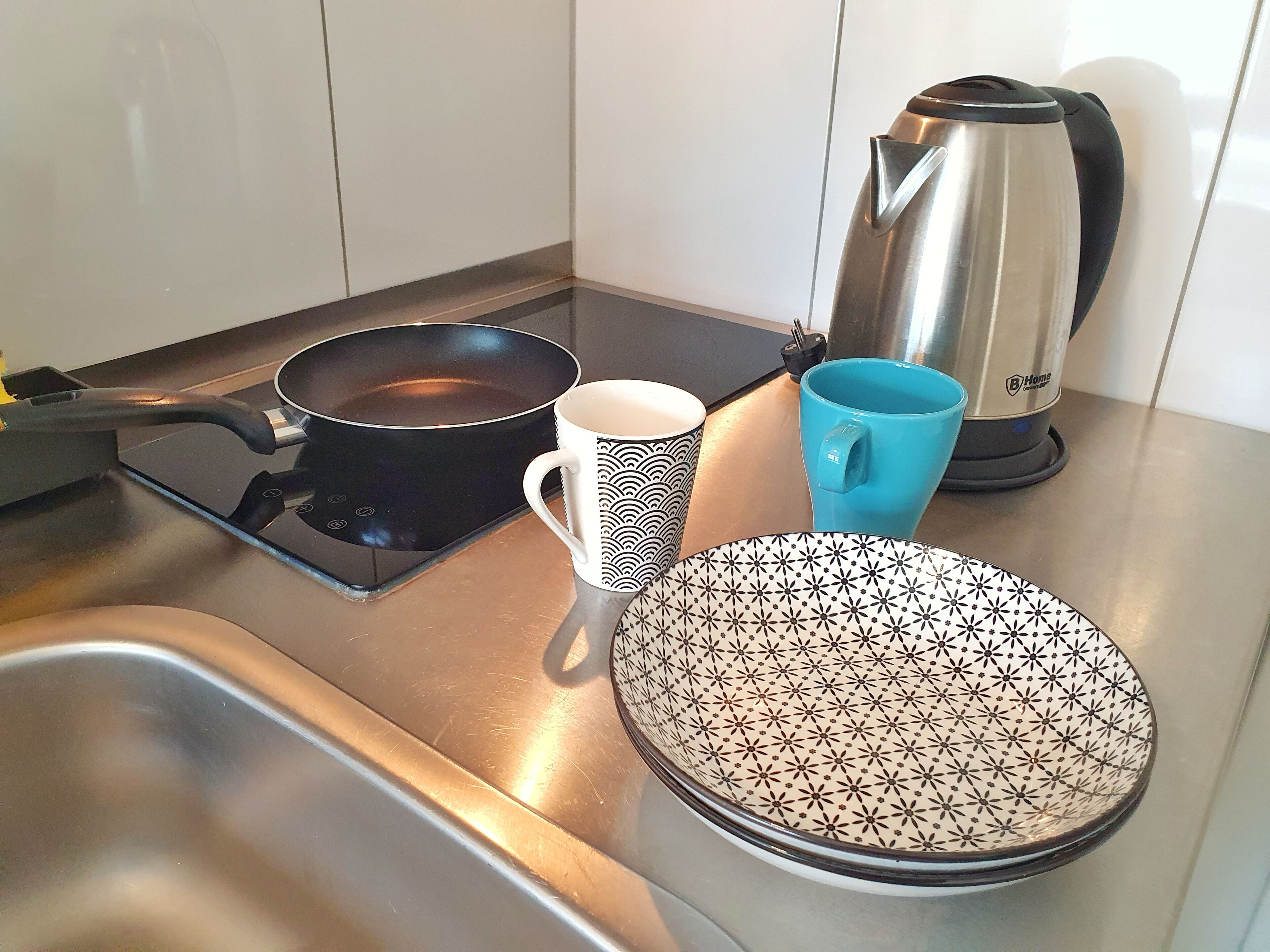 Ett kök med en spis, en stekpanna, en mugg, en blå mugg och en vattenkokare.