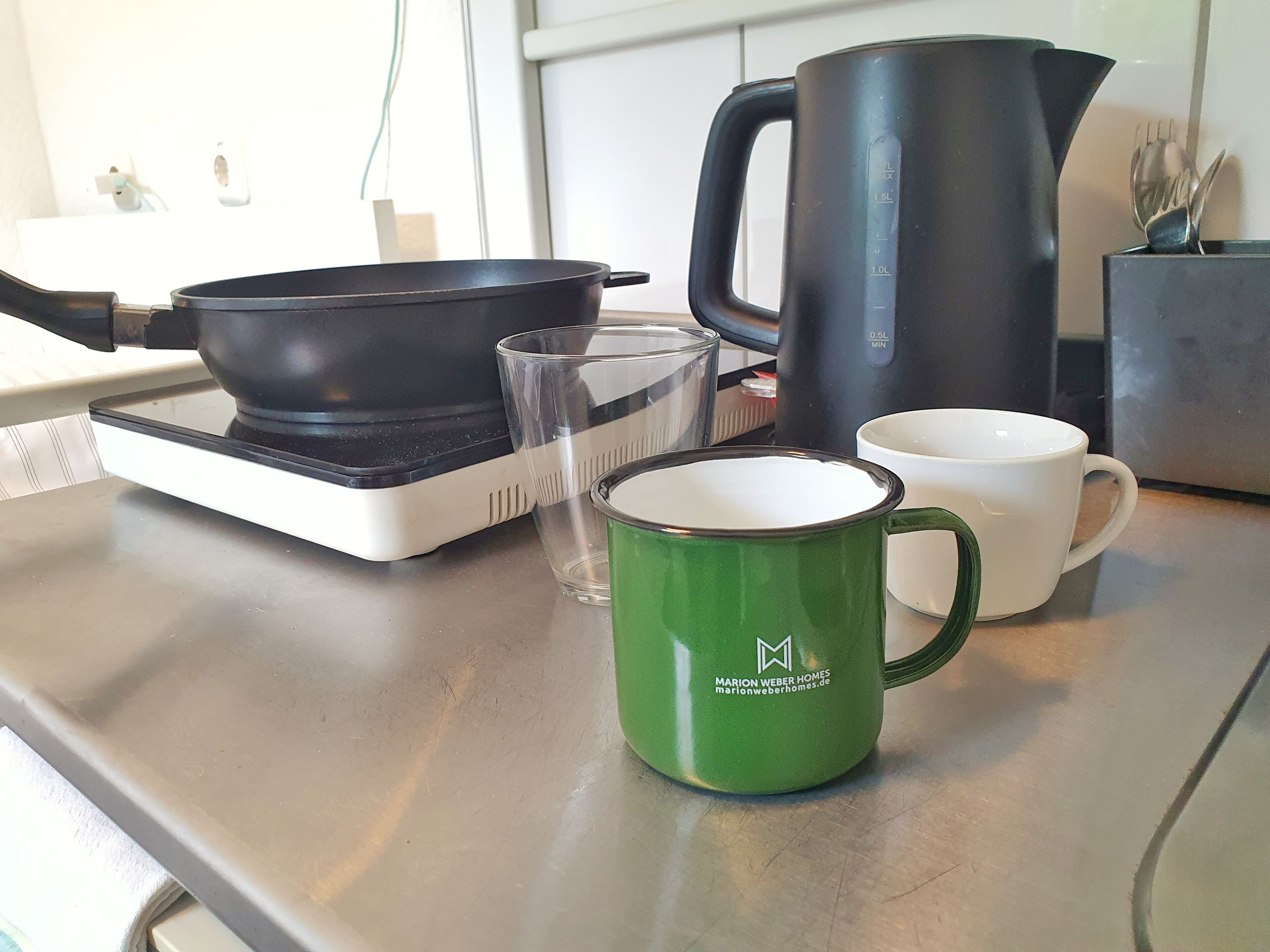 Una encimera de cocina con una sartén negra, un vaso, una taza verde y una taza blanca.