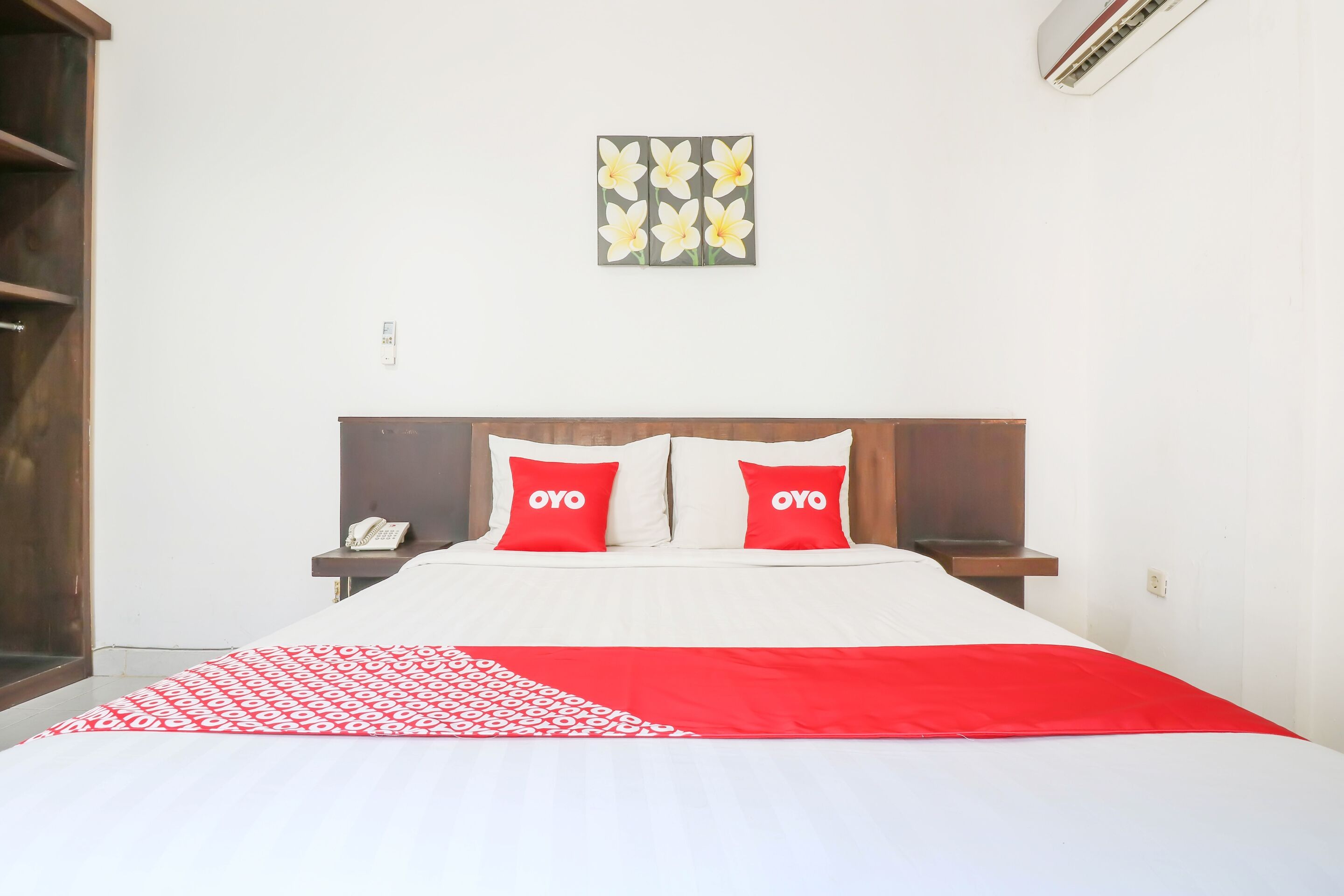 A neatly made bed with a red and white patterned bedspread, two red pillows with "OYO" text, a wall-mounted artwork of flowers, and a wooden nightstand with a phone.
