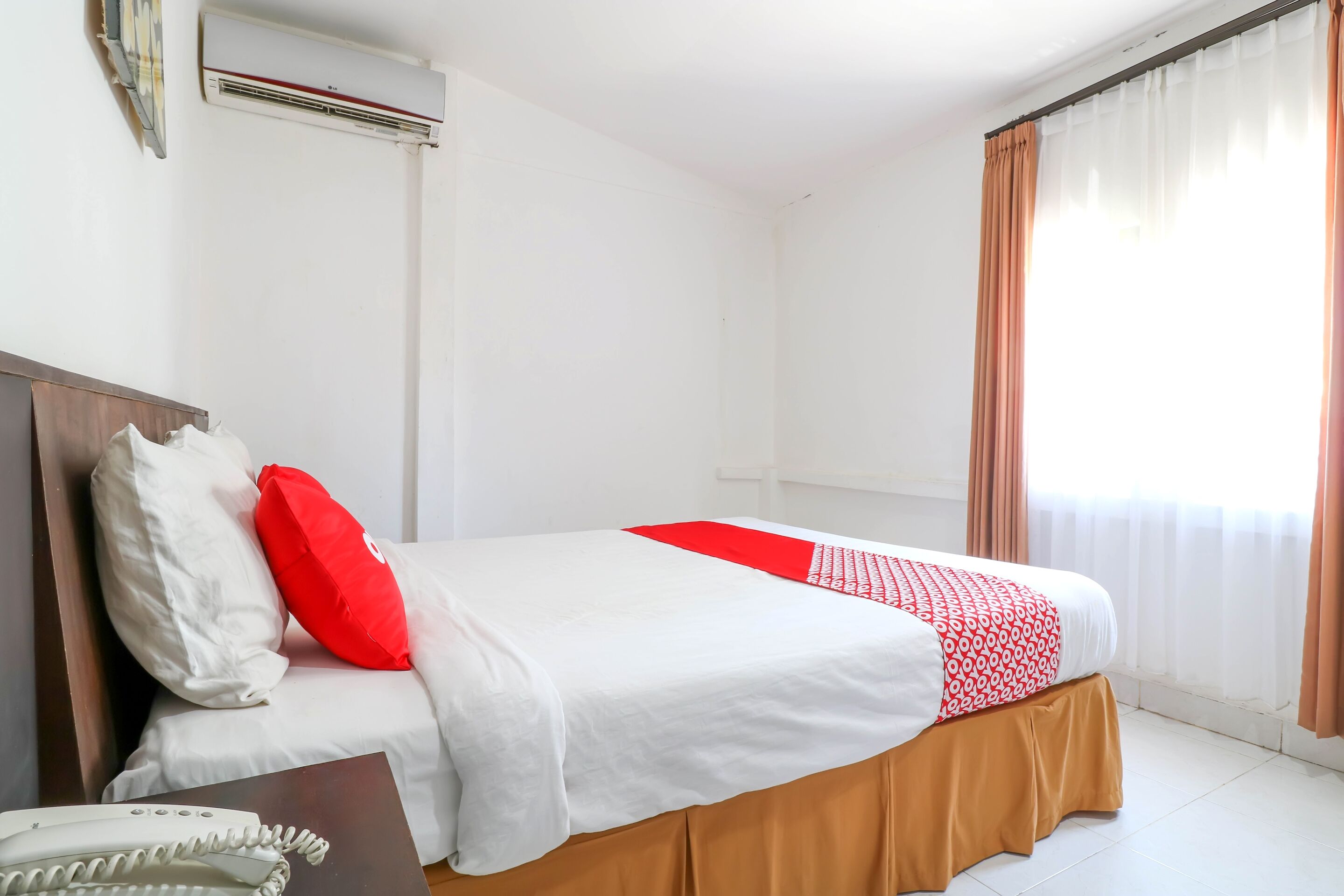 A hotel room with a bed, white walls, an air conditioning unit, a telephone, and a window with curtains.