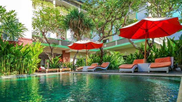 Un espace piscine avec des chaises longues et des parasols, entouré de verdure et avec un bâtiment en arrière-plan.