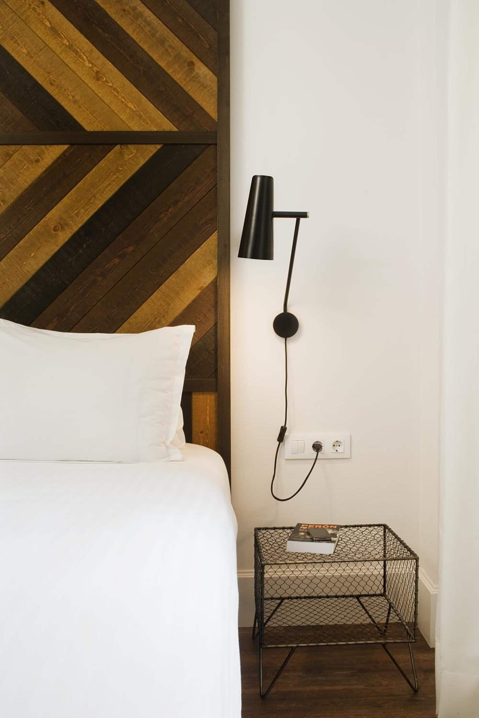 Un lit avec une literie blanche, une table de chevet en métal noir sur laquelle repose un livre, et une lampe fixée au mur.