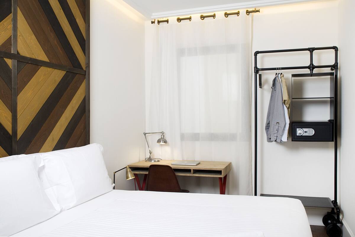 Une chambre d’hôtel comprenant un lit, un bureau avec une chaise, une lampe de bureau, un porte-manteau avec des vêtements et un mur orné d’un motif géométrique.