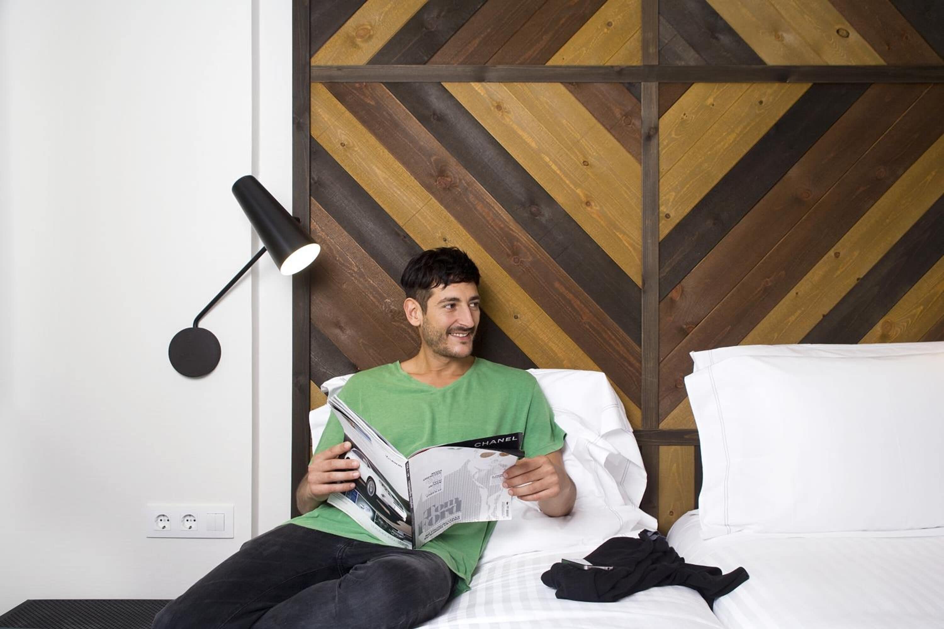 Una persona leyendo una revista en una cama en una habitación con un cabecero de madera con diseño en chevron y una lámpara de pie negra.
