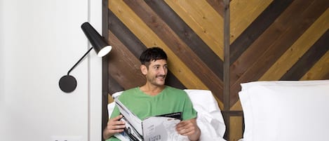 A person reading a magazine on a bed in a room with a wooden chevron-patterned headboard and a black floor lamp.