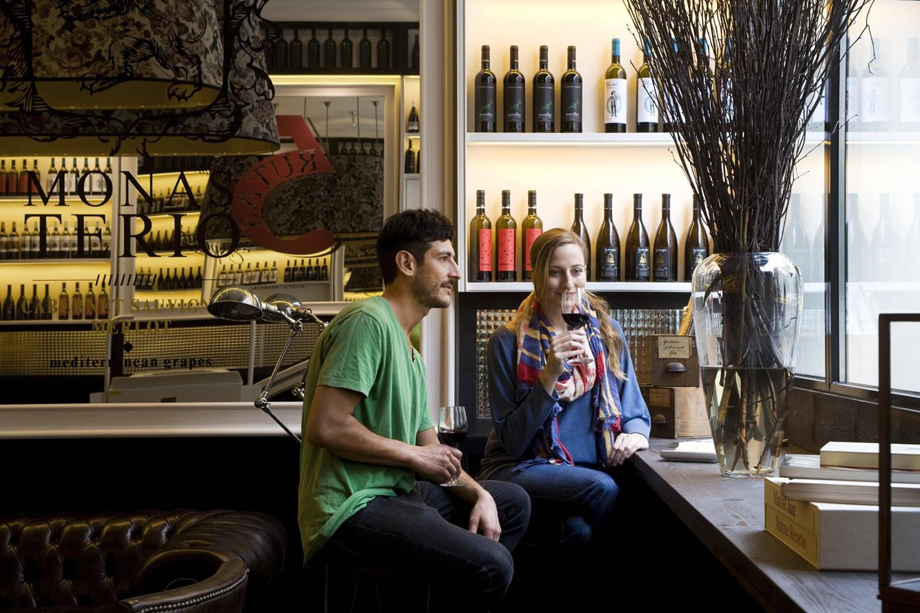 A bar with a variety of wine bottles on shelves, a decorative vase with branches, and a couple seated at a counter.
