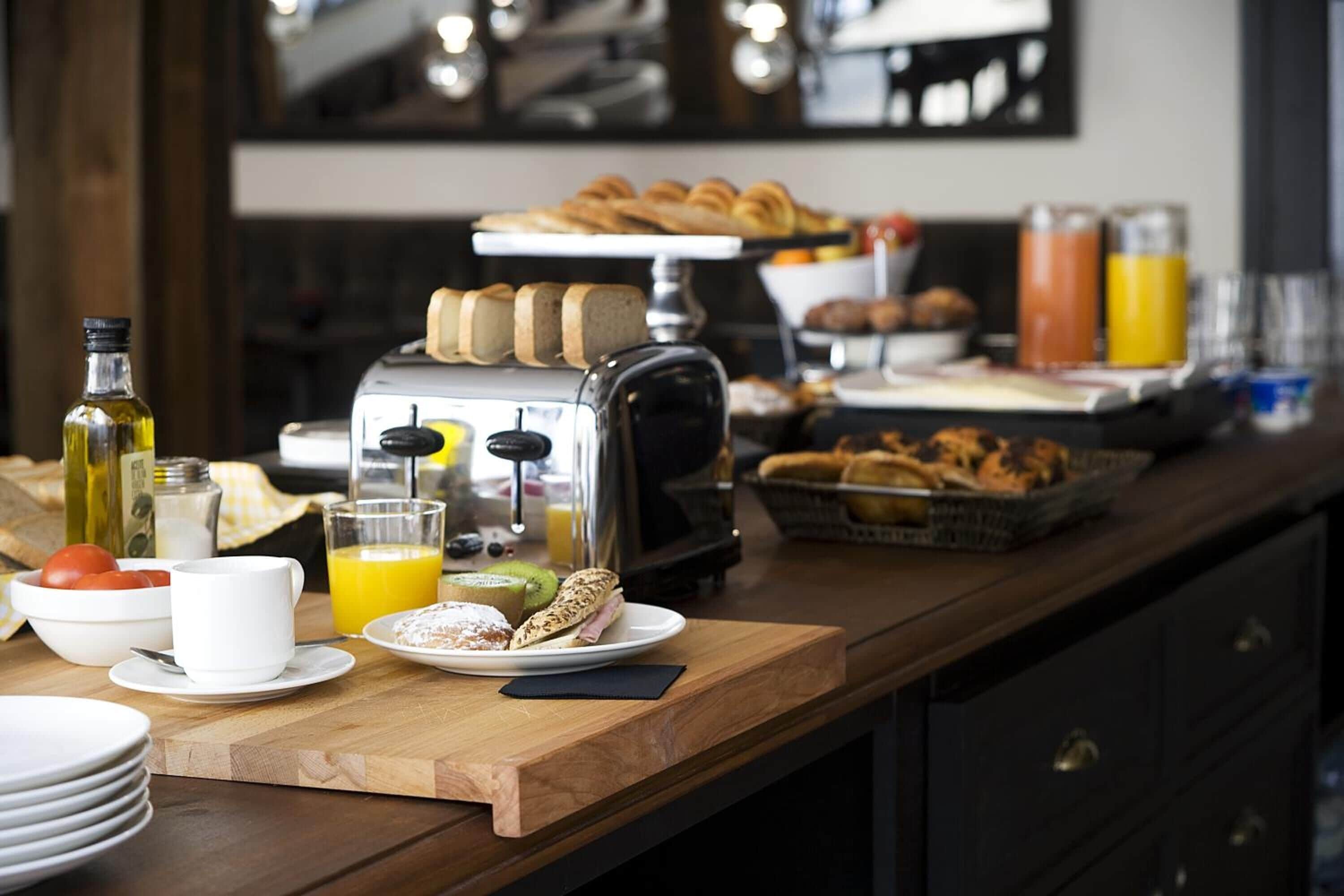 A breakfast buffet with various pastries, a toaster, and a juice dispenser on a wooden counter.