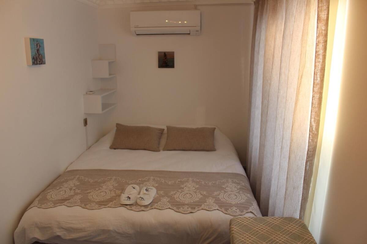 A small, neatly arranged bedroom with a bed, two pillows, a bedside table, and a pair of slippers.
