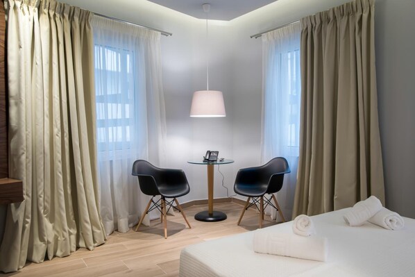 A hotel room with a bed, two black chairs, a small table with a lamp, and curtains on the windows.