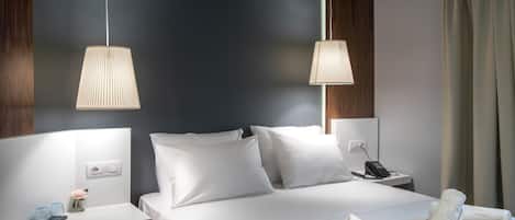 Una habitación de hotel moderna con una cama bien hecha, dos lámparas de noche, una mesita de noche con un teléfono y un pequeño estante con un florero y vasos.