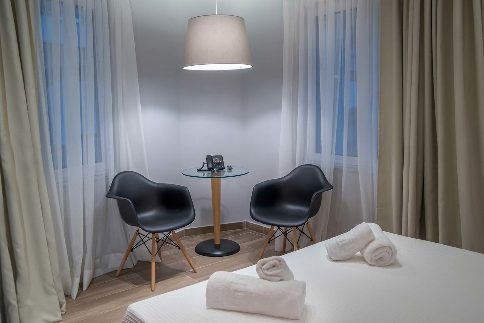 A hotel room with a bed, two black chairs, a small round table with a phone, and sheer curtains.