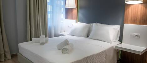 A neatly made bed with white linens and towels, a bedside table with a lamp, and a view of a building through sheer curtains.