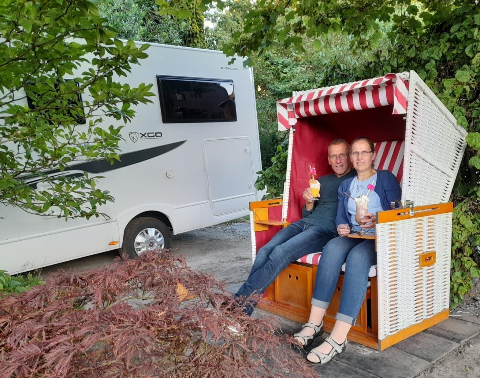 En vit husbil med "XCO"-loggan på sidan står parkerad bredvid en liten, randig strandstol.