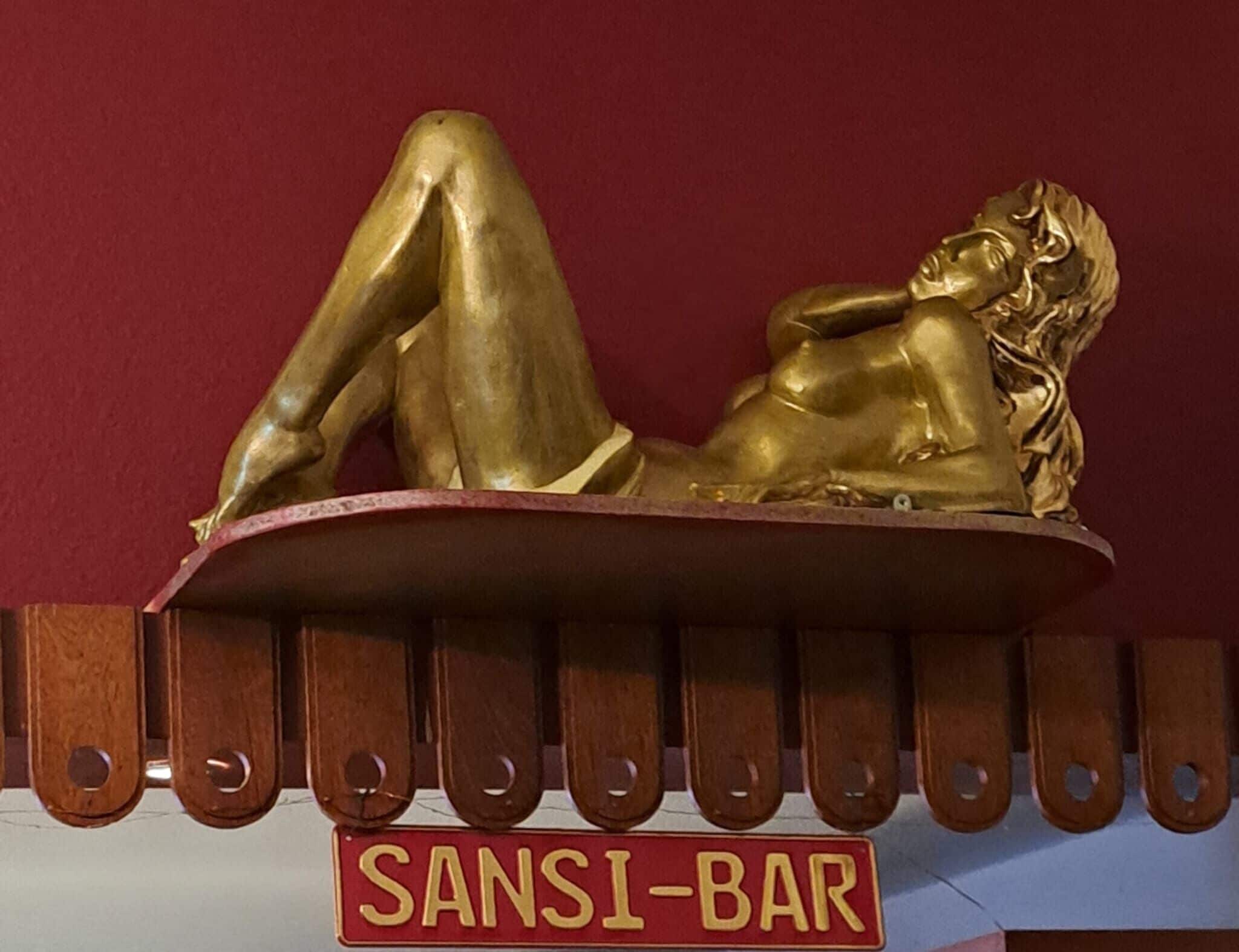 Uma estátua dourada de uma mulher deitada sobre uma plataforma, com uma placa indicando "SANSI-BAR" abaixo.