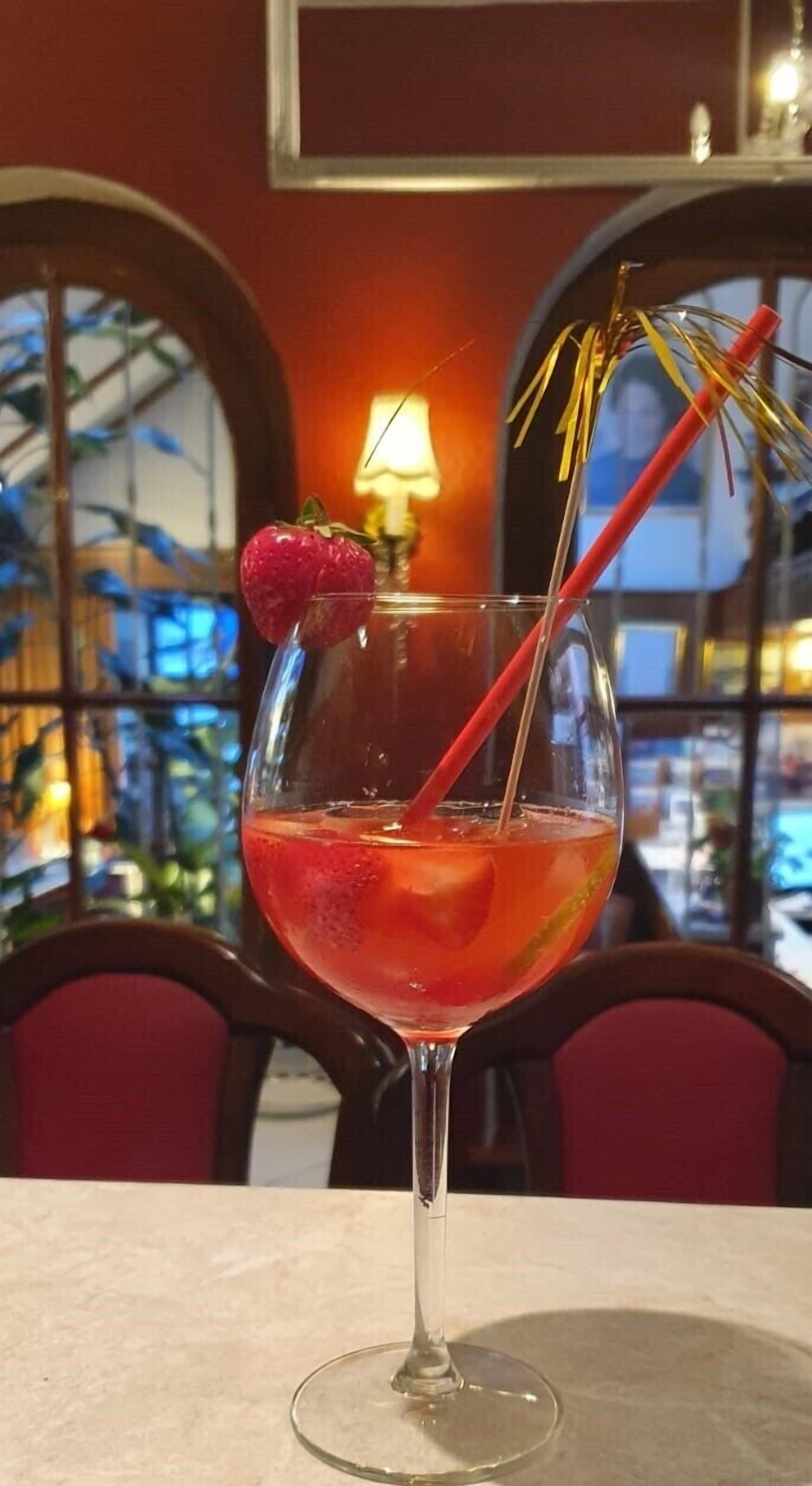A cocktail glass with a strawberry garnish on a table in a room with red chairs and a lamp in the background.
