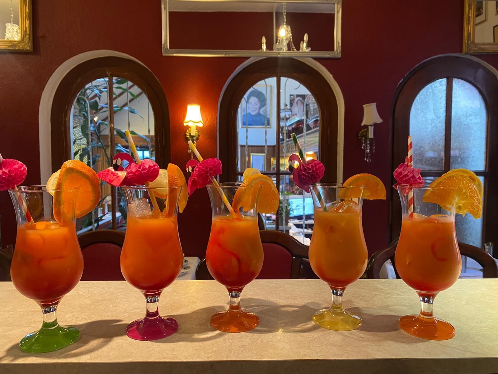 Una fila de coloridos cócteles con adornos, colocados sobre una mesa en una habitación con ventanales arqueados y un candelabro.