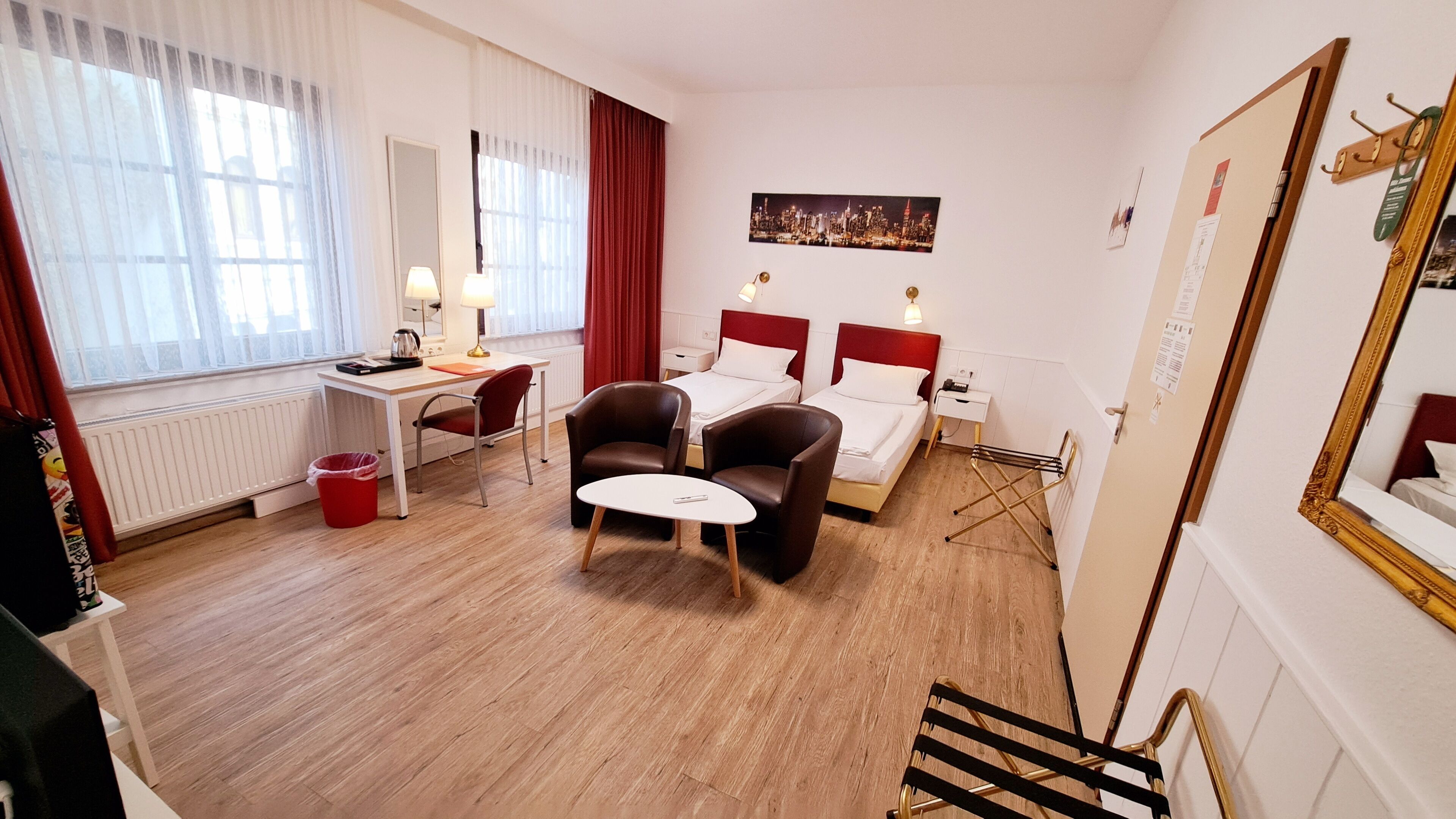 A hotel room with a wooden floor, a desk with a chair, two beds, a small table, and a window with curtains.