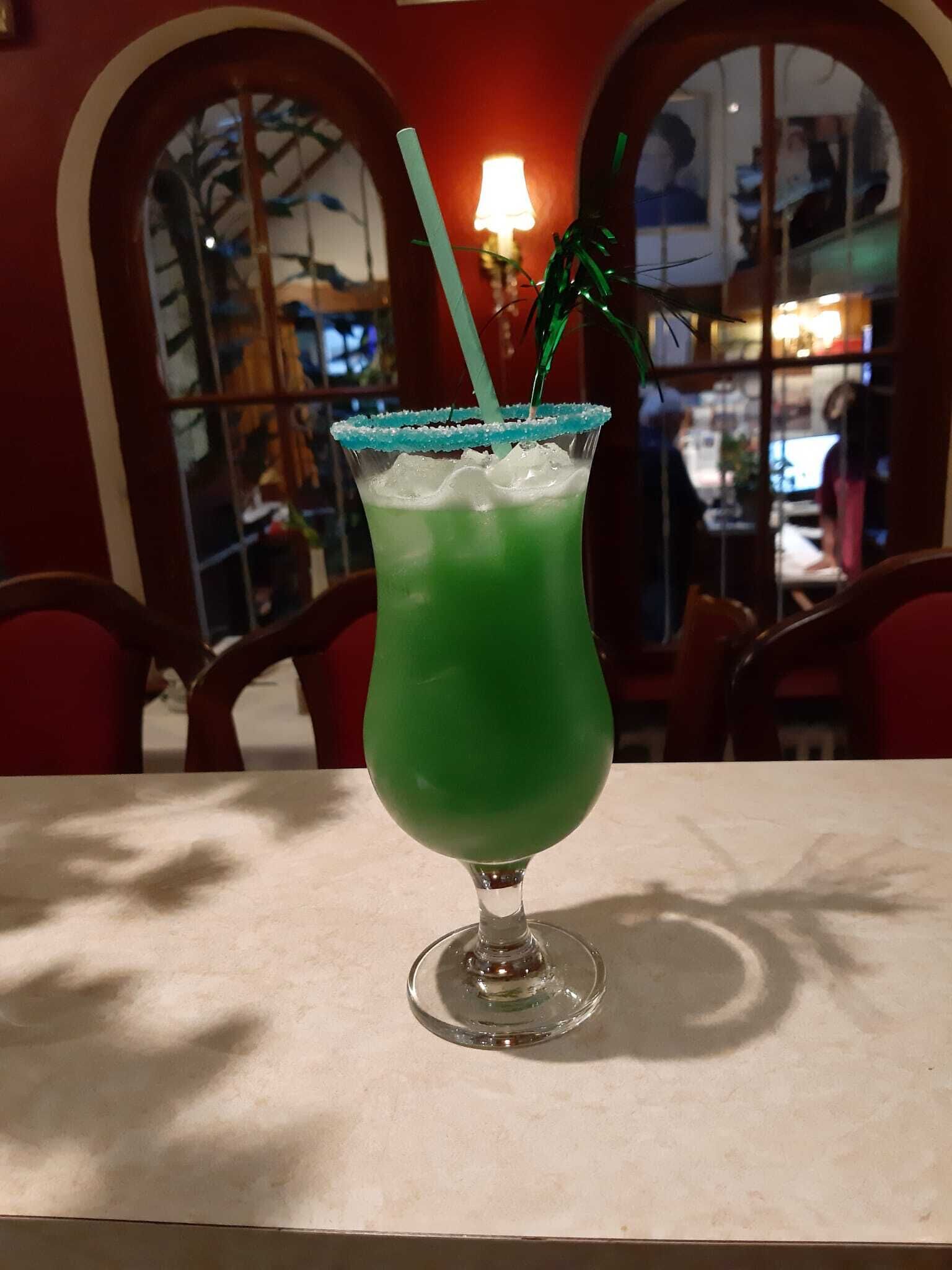 A green cocktail with a blue rim and a green straw in a glass, placed on a table in a room with arched windows and red walls.