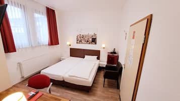 A hotel room with a double bed, a desk with a chair, a small table, a radiator, a window with curtains, and a cityscape picture on the wall.