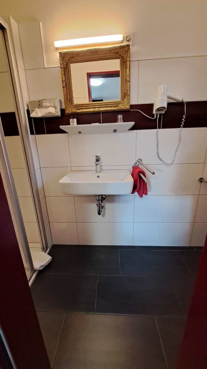 Un baño con un lavamanos blanco, un espejo con marco dorado, un secador de pelo y una toalla roja colgada en un perchero.