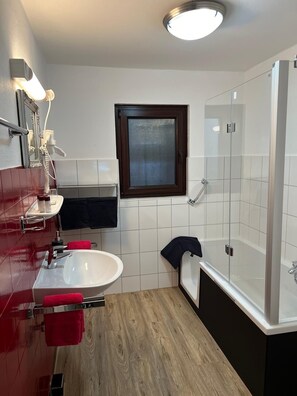 Un baño moderno con un lavamanos blanco, toallas rojas, cabina de ducha de vidrio y suelo de madera.