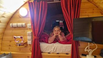 Un chalet confortable avec un intérieur en bois, un lit avec des rideaux rouges, une télévision et une petite étagère sur laquelle se trouve un objet en forme de chat.