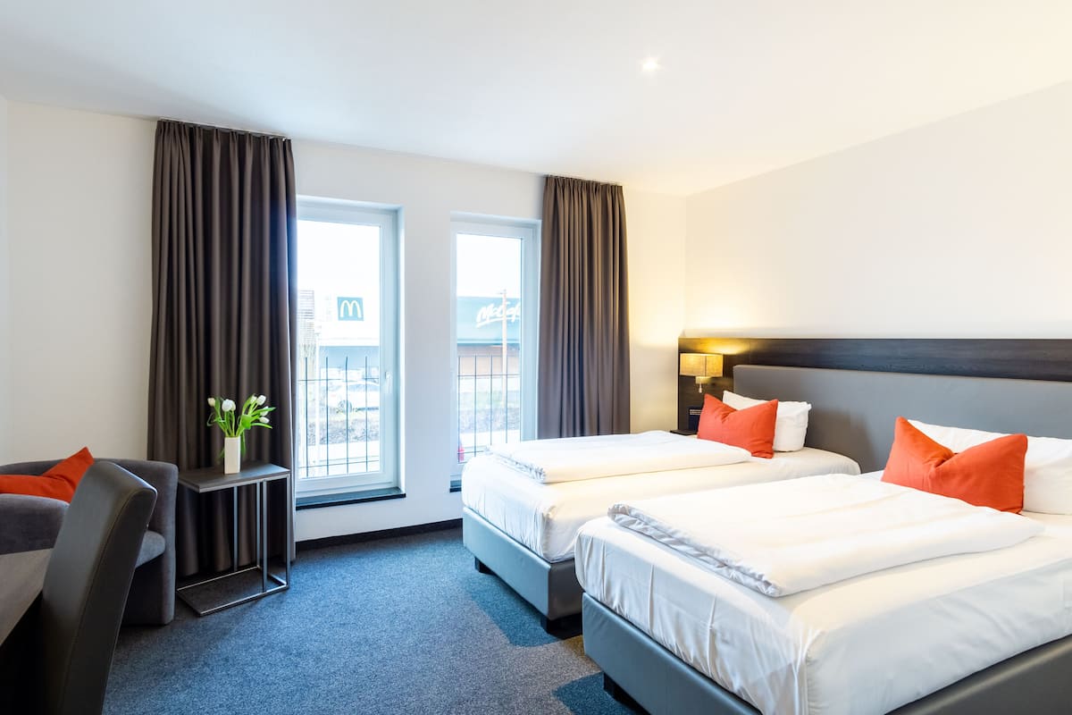 A hotel room with two beds, a desk, and a chair. There are curtains on the windows, and a plant on the desk.